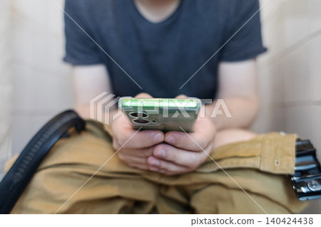 Man sitting on the toilet with a smartphone in hands. Guy scrolling phone on the loo, concept of prolonged sitting risk, screen addiction, and hemorrhoids. Man sitting on the toilet with a smartphone in hands. Guy scrolling phone on the loo, concept of prolonged sitting risk, screen addiction, and hemorrhoids. 140424438