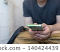 Man sitting on the toilet with a smartphone in hands. Guy scrolling phone on the loo, concept of prolonged sitting risk, screen addiction, and hemorrhoids. Man sitting on the toilet with a smartphone in hands. Guy scrolling phone on the loo, concept of prolonged sitting risk, screen addiction, and hemorrhoids. 140424439