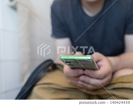 Man sitting on the toilet with a smartphone in hands. Guy scrolling phone on the loo, concept of prolonged sitting risk, screen addiction, and hemorrhoids. Man sitting on the toilet with a smartphone in hands. Guy scrolling phone on the loo, concept of prolonged sitting risk, screen addiction, and hemorrhoids. 140424439