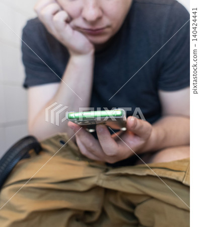 Man sitting on the toilet with a smartphone in hands. Guy scrolling phone on the loo, concept of prolonged sitting risk, screen addiction, and hemorrhoids. 140424441