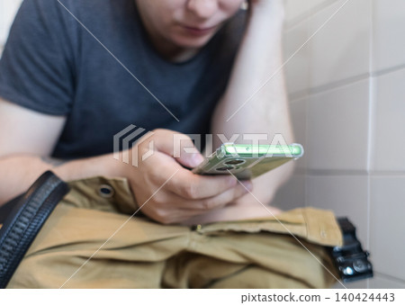 Man sitting on the toilet with a smartphone in hands. Guy scrolling phone on the loo, concept of prolonged sitting risk, screen addiction, and hemorrhoids. 140424443
