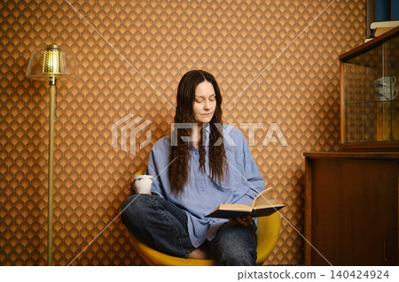 Woman reading in cozy chair with cup in hand in modern living room with unique wallpaper design Woman reading in cozy chair with cup in hand in modern living room with unique wallpaper design 140424924