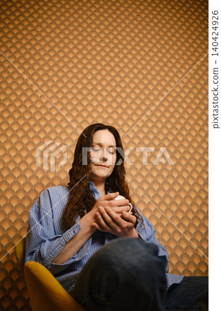 Woman sitting on chair holding cup in front of textured wall in indoor space Woman sitting on chair holding cup in front of textured wall in indoor space 140424926