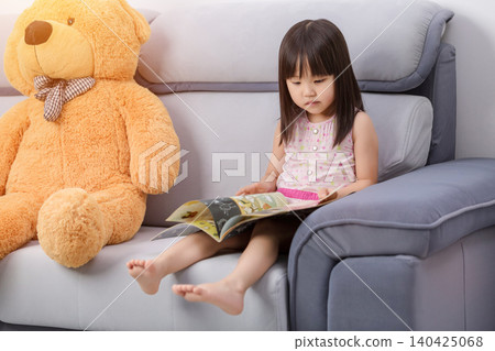 A giant teddy bear accompanies an Asian little girl as she reads on the sofa, a heartwarming scene of family learning and childhood ideals. A giant teddy bear accompanies an Asian little girl as she reads on the sofa, a heartwarming scene of family learning and childhood ideals. 140425068