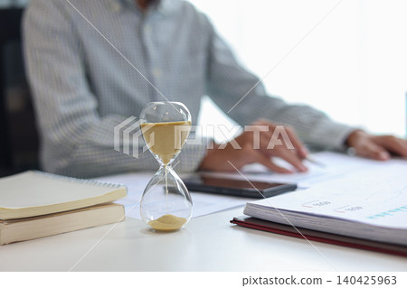 Businessman at a desk with an hourglass in the foreground, representing a time management concept. Businessman at a desk with an hourglass in the foreground, representing a time management concept. 140425963