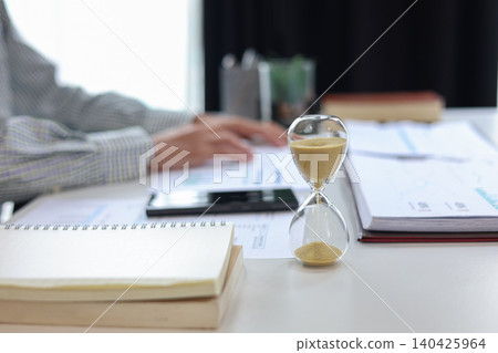 Businessman at a desk with an hourglass in the foreground, representing a time management concept. Businessman at a desk with an hourglass in the foreground, representing a time management concept. 140425964