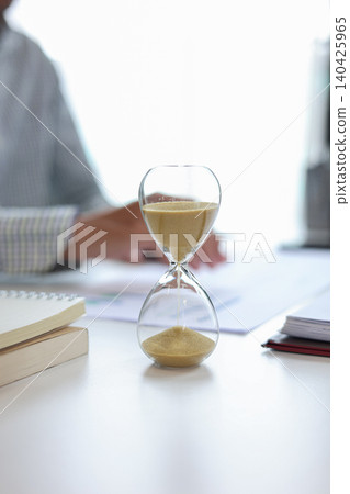 Businessman at a desk with an hourglass in the foreground, representing a time management concept. Businessman at a desk with an hourglass in the foreground, representing a time management concept. 140425965