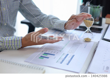 Professional analyzing company financial report and budget graphs at desk. Time management and financial analysis concept with hourglass on desk. Professional analyzing company financial report and budget graphs at desk. Time management and financial analysis concept with hourglass on desk. 140425986