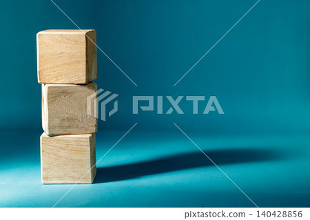 Three blank wooden cubes stacked vertically on a dark blue background with a strong light source casting a long, dramatic shadow to the right. High Quality Photo Three blank wooden cubes stacked vertically on a dark blue background with a strong light source casting a long, dramatic shadow to the right. High Quality Photo 140428856