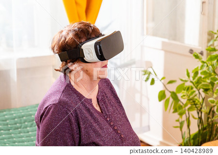 Elderly user experiencing virtual reality simulation with VR headset in a comfortable home setting. Digital inclusion and innovation concept. Aging with technology and curiosity. Close up Elderly user experiencing virtual reality simulation with VR headset in a comfortable home setting. Digital inclusion and innovation concept. Aging with technology and curiosity. Close up 140428989