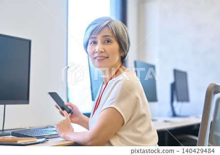 一位頭髮花白的女士在辦公桌前轉過身，手裡拿著她的智慧型手機。 140429484