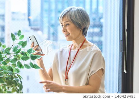 一位頭髮花白的女士靠在窗邊,一邊看著她的智慧型手機,一邊微笑。 一位頭髮花白的女士靠在窗邊,一邊看著她的智慧型手機,一邊微笑。 140429485
