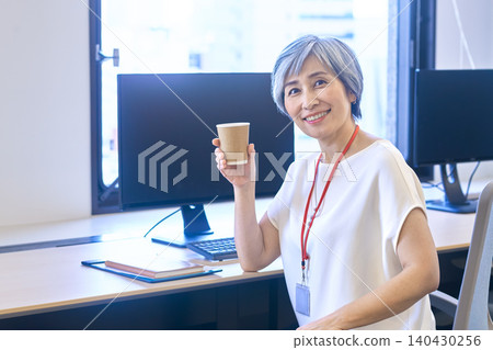 A gray-haired woman smiles, holding a paper cup at her office desk. A gray-haired woman smiles, holding a paper cup at her office desk. 140430256