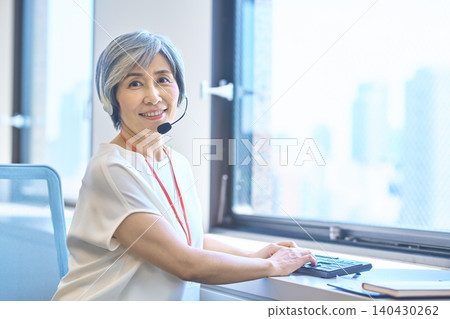 一位頭髮花白、戴著耳機的女士從靠窗的桌子旁轉過身來。 一位頭髮花白、戴著耳機的女士從靠窗的桌子旁轉過身來。 140430262