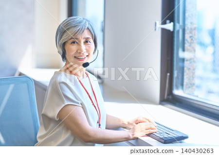 A gray-haired woman working with a smile while wearing a headset. A gray-haired woman working with a smile while wearing a headset. 140430263