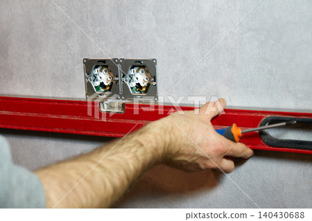 Electrician aligns and installs wall power outlets using a spirit level and screwdriver during interior renovation. Electrician aligns and installs wall power outlets using a spirit level and screwdriver during interior renovation. 140430688