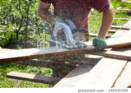 Worker using a circular saw to cut wooden boards outdoors, with sawdust flying as part of construction  140430769