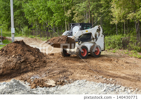 Skid steer loader carrying a bucket of soil along a dirt road at a construction site Skid steer loader carrying a bucket of soil along a dirt road at a construction site 140430771