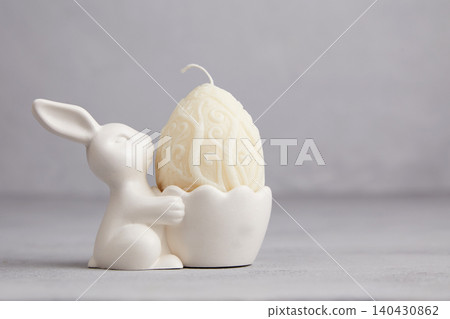 A white Easter egg-shaped candle in a bunny-shaped holder against a gray background A white Easter egg-shaped candle in a bunny-shaped holder against a gray background 140430862