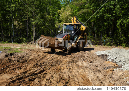 Backhoe loader carrying and dumping soil at a construction site, shaping ground and preparing foundation area 140430878