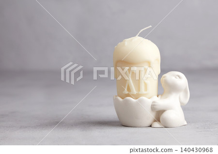 A white candle shaped like an Easter cake sits in a bunny-shaped candlestick against a gray background. A white candle shaped like an Easter cake sits in a bunny-shaped candlestick against a gray background. 140430968
