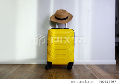 A yellow suitcase with a straw hat sits against the wall of a hotel room or apartment. A yellow suitcase with a straw hat sits against the wall of a hotel room or apartment. 140431167