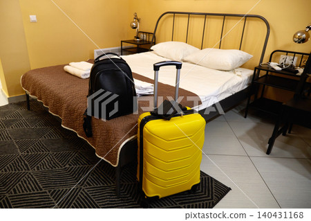 Bright yellow suitcase and black carry-on beside a bed in a hotel room with neatly folded towels 140431168