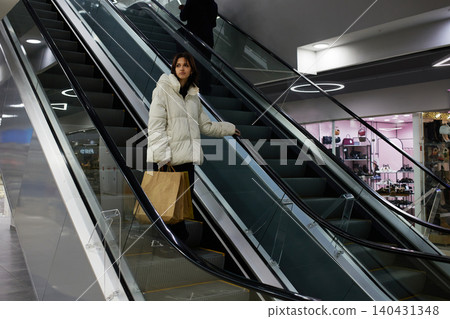 Woman wearing a white jacket stands on an escalator in a shopping mall while holding a shopping bag Woman wearing a white jacket stands on an escalator in a shopping mall while holding a shopping bag 140431348