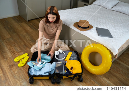 A woman puts hygiene products, clothes, a travel guide, headphones and travel accessories in a suitcase A woman puts hygiene products, clothes, a travel guide, headphones and travel accessories in a suitcase 140431354