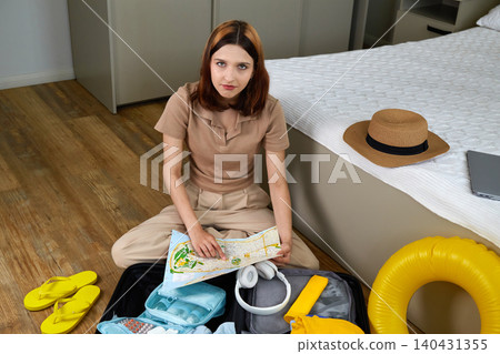 The girl packs her suitcase for a trip. She studies the map and plans the route of the trip The girl packs her suitcase for a trip. She studies the map and plans the route of the trip 140431355