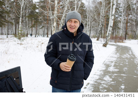 A happy man walks through a snowy forest in winter, holding a cup of hot drink in his hands A happy man walks through a snowy forest in winter, holding a cup of hot drink in his hands 140431451