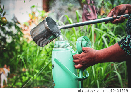 Weathered hands pouring steaming hot water from metal ladle into mint green thermos flask in lush garden Weathered hands pouring steaming hot water from metal ladle into mint green thermos flask in lush garden 140431569