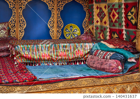 Traditional uzbek interior with colorful ikat silk fabric and ornate carved wooden furniture. Ethnic central asian style room decoration with handmade textiles and decorative plates. Traditional uzbek interior with colorful ikat silk fabric and ornate carved wooden furniture. Ethnic central asian style room decoration with handmade textiles and decorative plates. 140431637