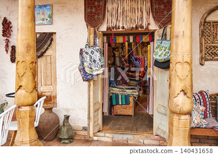 Entrance to traditional souvenir shop with colorful handmade bags and oriental fabrics. Exterior of ethnic handicraft store with wooden carved columns. Cultural tourism and local craft. Uzbekistan. Entrance to traditional souvenir shop with colorful handmade bags and oriental fabrics. Exterior of ethnic handicraft store with wooden carved columns. Cultural tourism and local craft. Uzbekistan. 140431658