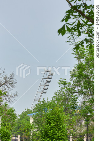 Metal stadium floodlight tower rising above green trees. Outdoor sports arena illumination infrastructure. Tall industrial metal construction for night event lighting. Metal stadium floodlight tower rising above green trees. Outdoor sports arena illumination infrastructure. Tall industrial metal construction for night event lighting. 140431659