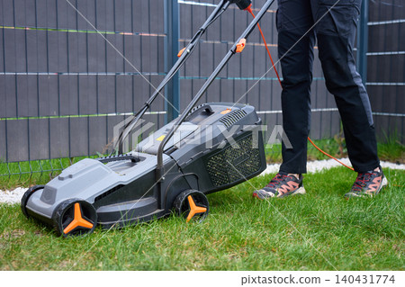 Lawn mower with grass collector and power cord stands on backyard grass near person Lawn mower with grass collector and power cord stands on backyard grass near person 140431774