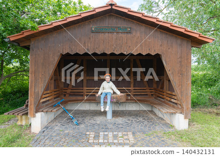 A cute little boy with a denim backpack sitting on a wooden bench inside a rustic rural bus stop shelter. Perfect commercial concept for childhood journeys, countryside travel, and summer holidays A cute little boy with a denim backpack sitting on a wooden bench inside a rustic rural bus stop shelter. Perfect commercial concept for childhood journeys, countryside travel, and summer holidays 140432131