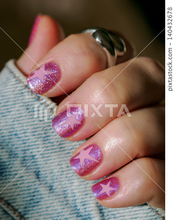 Pink star nail art manicure with denim jacket. Female hand showing sparkling pink and blue shimmer nails with star design next to blue jeans fabric. Modern youth fashion and beauty salon concept 140432678
