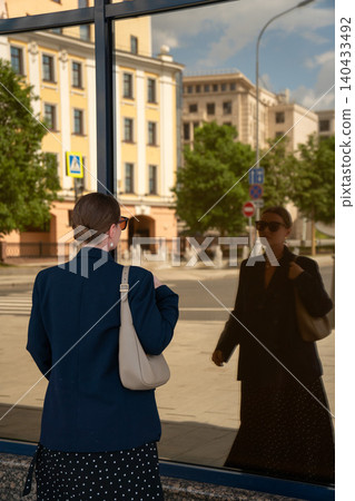 Woman observes her reflection in glass facade in Moscow, historic urban architecture mirrored in modern surface, identity, self-perception, contemporary city life. Fashion content, confidence concept Woman observes her reflection in glass facade in Moscow, historic urban architecture mirrored in modern surface, identity, self-perception, contemporary city life. Fashion content, confidence concept 140433492