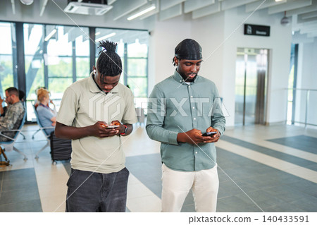 Two young adult men using mobile phones at airport lounge Two young adult men using mobile phones at airport lounge 140433591