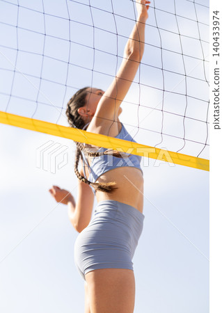 Young woman spiking a volleyball during a match on a sunny day at the beach with blue sky and net in the background 140433974