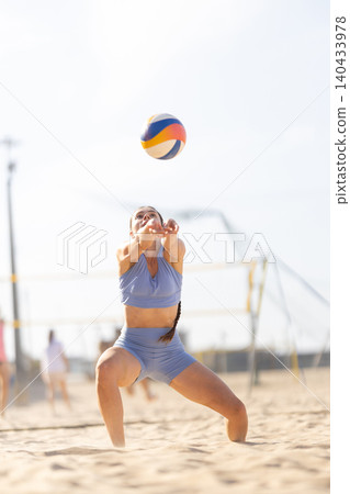 Woman plays beach volleyball, receiving a ball during a match at a sandy court under a bright sun near a sports facility Woman plays beach volleyball, receiving a ball during a match at a sandy court under a bright sun near a sports facility 140433978