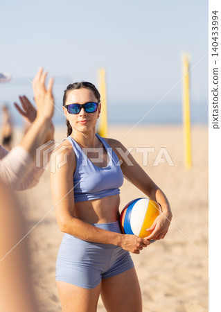 Young woman holding beach volleyball on sandy shore with sun shining and players in the background during summer day Young woman holding beach volleyball on sandy shore with sun shining and players in the background during summer day 140433994