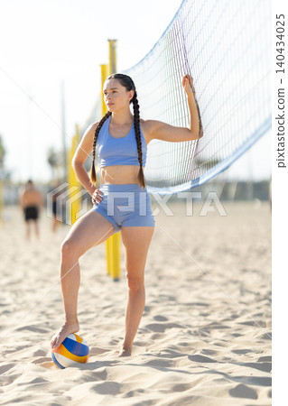 Young woman poses by volleyball net on beach during sunny day while others play in background Young woman poses by volleyball net on beach during sunny day while others play in background 140434025