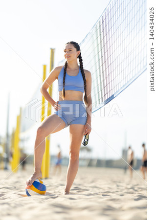 Young woman stands on the beach with a volleyball while enjoying a sunny day with friends playing sports near the net Young woman stands on the beach with a volleyball while enjoying a sunny day with friends playing sports near the net 140434026