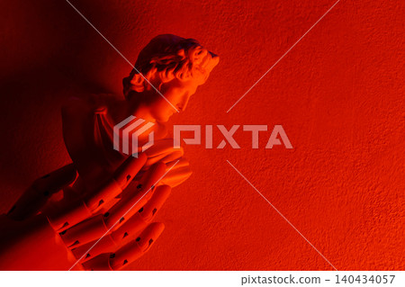 Classical angel statue with praying hands illuminated by dramatic red lighting on textured wall 140434057