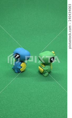 Two small cute blue and green toy dinosaur figures standing on bright green background. Concept of childhood fun Two small cute blue and green toy dinosaur figures standing on bright green background. Concept of childhood fun 140434063
