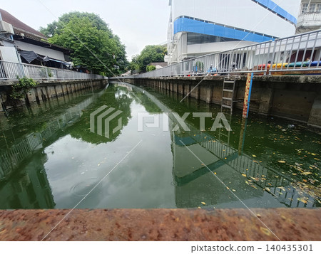 Urban canal waterway, concrete bank, calm reflection, green water, drainage channel, city landscape Urban canal waterway, concrete bank, calm reflection, green water, drainage channel, city landscape 140435301