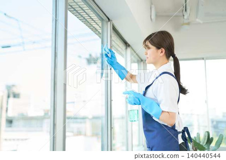 A young woman in an apron carefully wipes a window. A young woman in an apron carefully wipes a window. 140440544