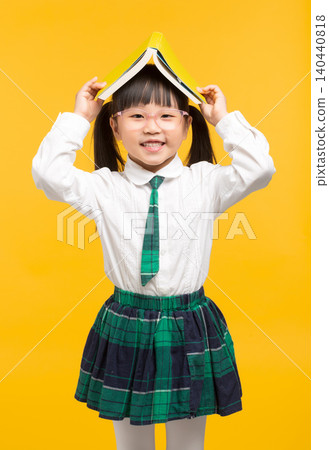 A playful elementary school girl balances a yellow textbook on her head with her hands up, showing a fun side of school life. A playful elementary school girl balances a yellow textbook on her head with her hands up, showing a fun side of school life. 140440818